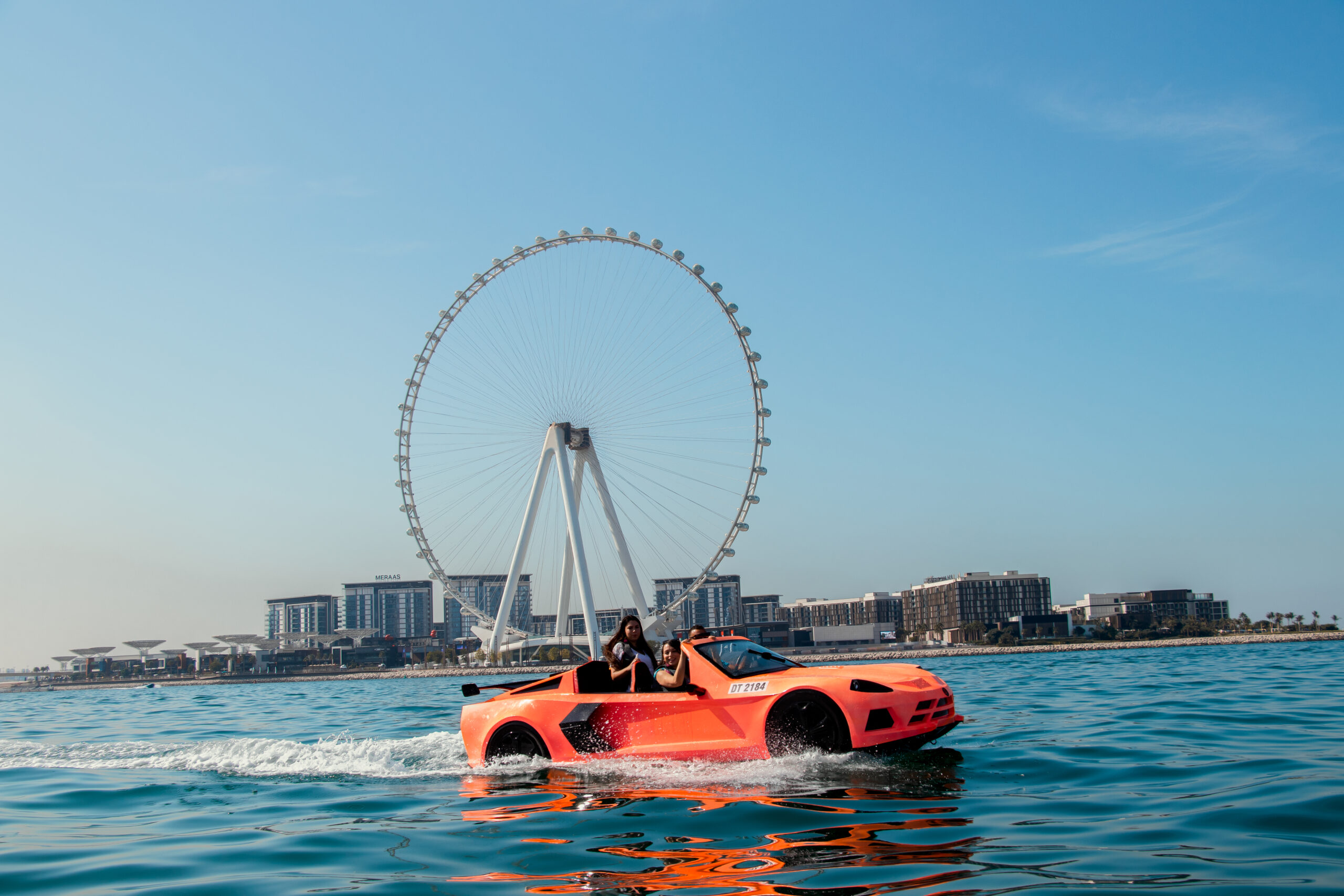 Two people in an orange amphibious jet car on the water with a giant observation wheel in the background.